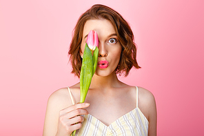 young woman in white dress covering eye with pink flower isolated on pink