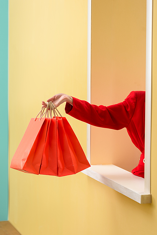 partial view of stylish woman in red clothing with red shopping bags outstretching hand out decorative window