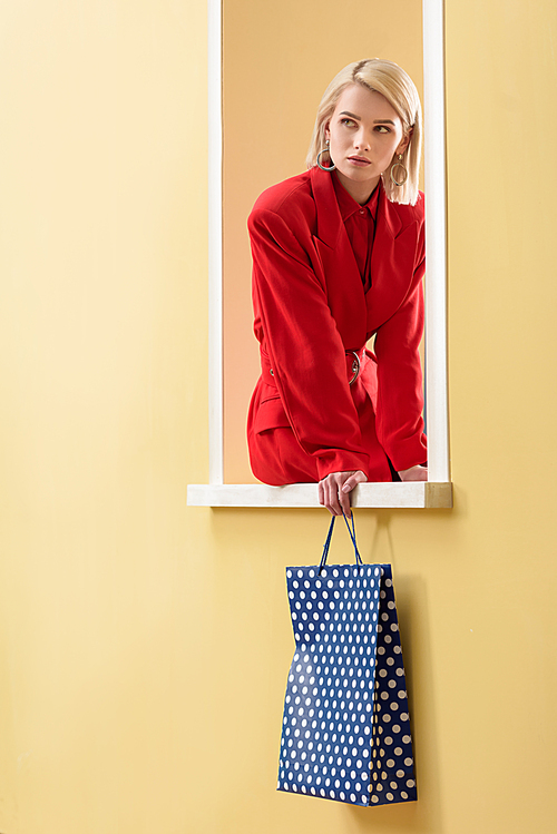 stylish pensive woman in red suit with shopping bag with dots pattern in hand sitting on decorative window