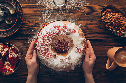 cropped view of female hands holding traditional homemade christmas cake with pomegranate and nuts and wooden tabletop