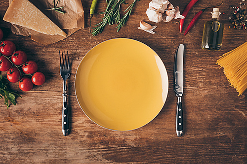 top view of row pasta, fresh ingredients and plate with knife and fork on wooden tabletop