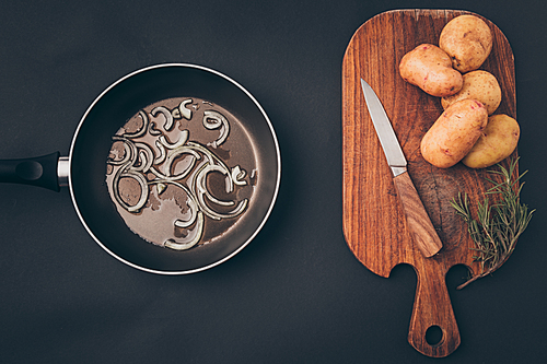 top view of frying pan with onion and wooden board with potatoes on gray surface