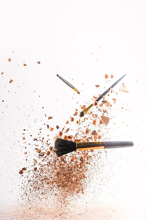 crushed cosmetic powder with makeup brushes falling isolated on white