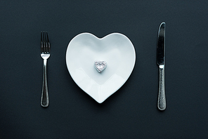 top view of wrapped candy on white heart shaped plate with cutlery isolated on black