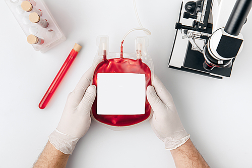 top view of hands in gloves holding blood for transfusion near vials and microscope isolated on white