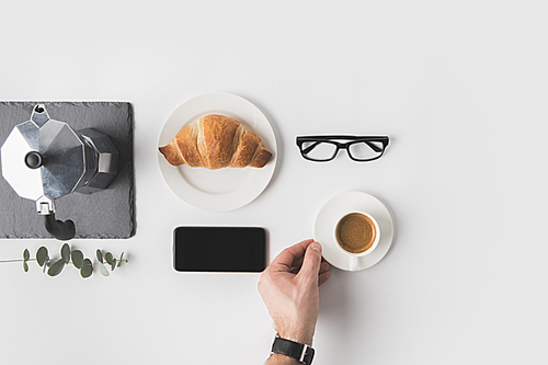 cropped shot of man holding cup of coffee with smartphone, eyeglasses and croissant for breakfast near by on white surface