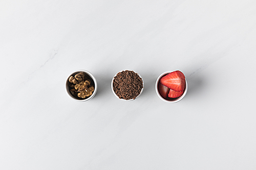 Three bowls with walnuts, grated chocolate and strawberries on white