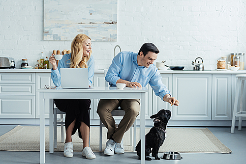 Man and woman drinking coffee and feeding french bulldog by kitchen table