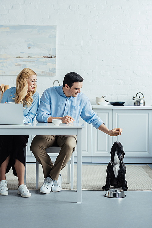Man and woman drinking coffee and playing with their dog by kitchen table with laptop