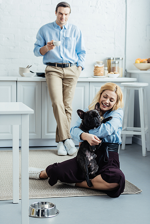 Man drinking coffee and blonde woman hugging cute frenchie on kitchen floor