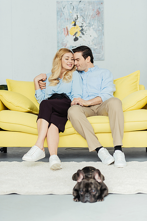 Blonde woman hugging her boyfriend on sofa by french bulldog on floor