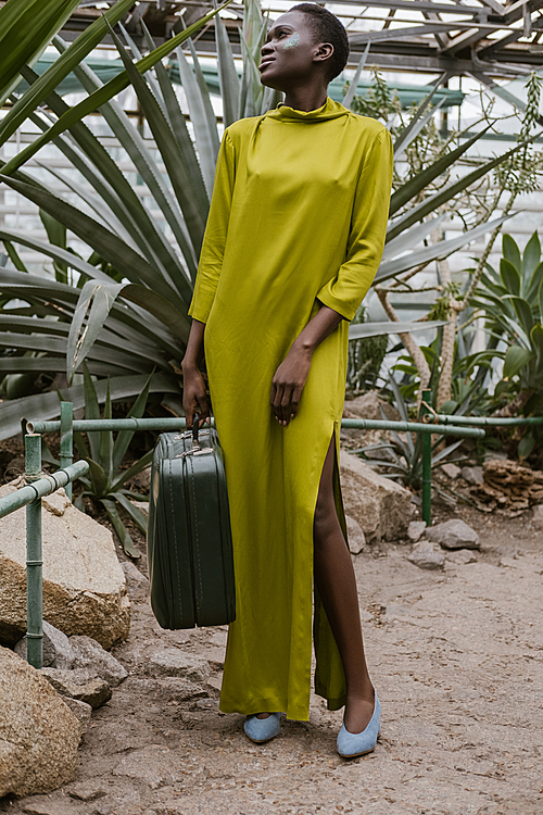 stylish african american woman in yellow dress posing with travel bag in tropical greenhouse