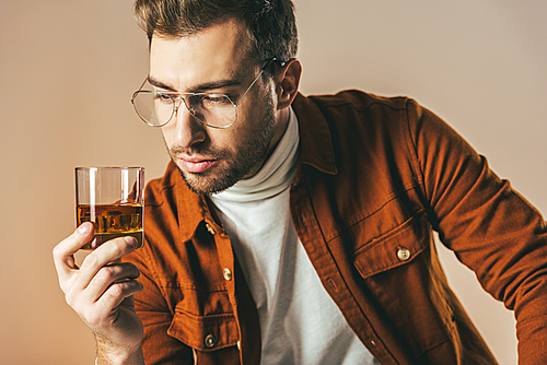 portrait of thoughtful fashionable man looking at glass of cognac in hand isolated on beige