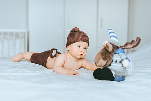 infant child in knitted deer shorts and hat playing with toy moose in bed