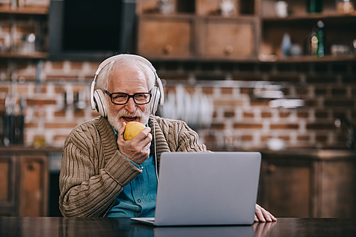 Happy senior man in headphones eating apple using laptop