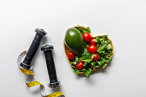 top view of vegetables in heart-shaped bowl, measuring tape and dumbbells on white background