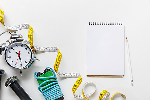 top view of sport equipment, measuring tape, alarm clock near blank notebook on white background