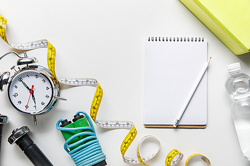 top view of sport equipment, measuring tape, alarm clock, water and blank notebook on white background