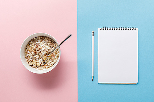 top view of breakfast cereal in bowl and blank notebook with pencil on blue and pink background