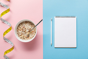 top view of breakfast cereal in bowl near measuring tape and blank notebook with pencil on blue and pink background
