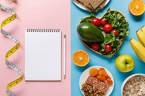 top view of diet food on blue and empty notebook with pencil and measuring tape on pink background