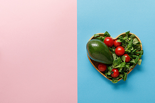 top view of fresh green vegetable salad in heart shaped bowl on pink and blue background