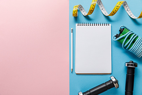 top view of blank notebook, dumbbells, skipping rope and measuring tape on blue and pink background