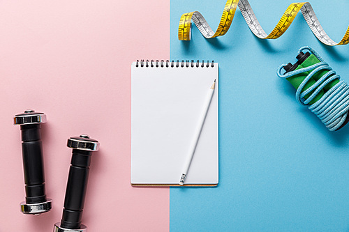 top view of empty notebook, dumbbells, skipping rope and measuring tape on blue and pink background