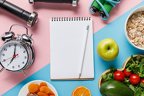 top view of empty notebook and pencil with delicious diet food and sport equipment on blue and pink background