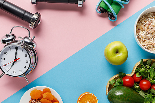 top view of delicious diet food and sport equipment with alarm clock on blue and pink background