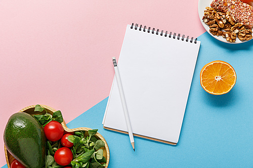 top view of empty notebook, vegetable salad, orange and nuts on pink and blue background