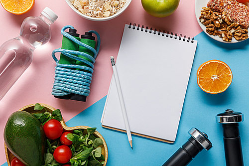 top view of empty notebook and pencil with delicious diet food, water and sport equipment on blue and pink background
