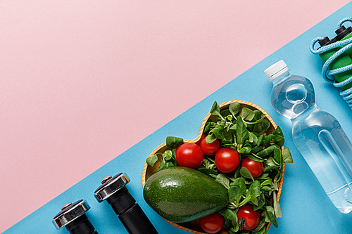 flat lay with vegetable salad in heart-shaped bowl, water, skipping rope and dumbbells on pink and blue background