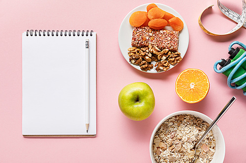 top view of fresh diet food, measuring tape, skipping rope and blank notebook on pink background with copy space