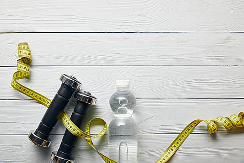top view of bottle with water, yellow measuring tape and dumbbells on wooden white background with copy space