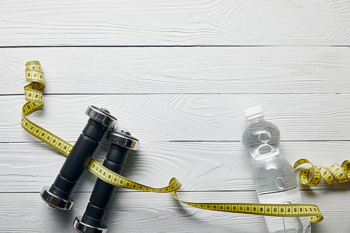 top view of bottle with water, measuring tape and dumbbells on wooden white background with copy space