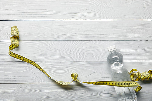 top view of transparent bottle with water and yellow measuring tape on wooden white background with copy space