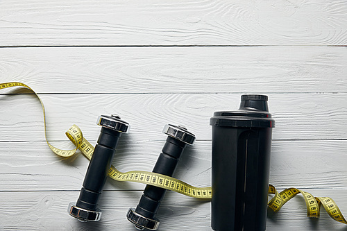 top view of black shaker cup, measuring tape and dumbbells on wooden white background with copy space
