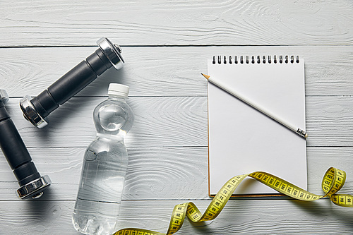 top view of dumbbells, water in bottle, measuring tape and blank notebook with copy space and pencil on wooden white background