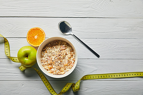 top view of measuring tape, spoon and breakfast cereal in bowl near apple and orange on wooden white background