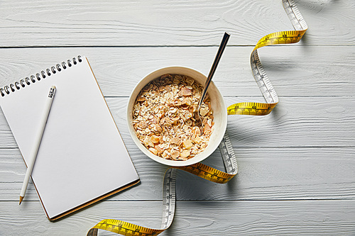 top view of measuring tape, spoon and breakfast cereal in bowl near notebook and pencil on wooden white background