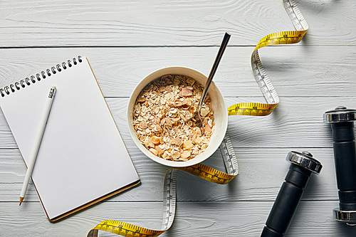 top view of measuring tape, spoon and breakfast cereal in bowl near notebook, dumbbells and pencil on wooden white background