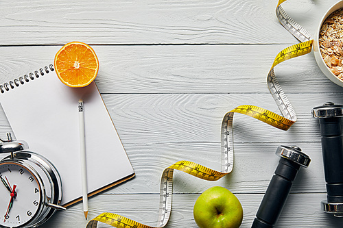 top view of measuring tape, breakfast cereal in bowl near apple, orange, notebook, dumbbells, alarm clock and pencil on wooden white background with copy space