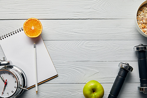 top view of breakfast cereal in bowl near apple, orange, notebook, dumbbells, alarm clock and pencil on wooden white background with copy space