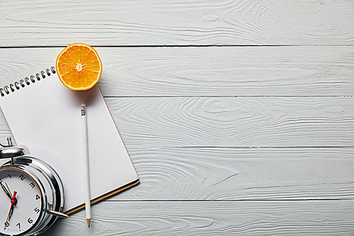 top view of orange half, notebook and pencil near alarm clock on wooden white background with copy space