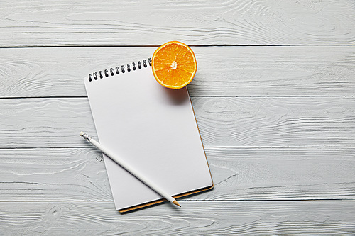 top view of orange half, blank notebook and pencil on wooden white background with copy space