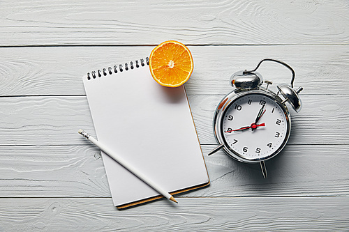 top view of orange half, blank notebook and pencil near alarm clock on wooden white background