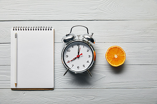 flat lay with silver alarm clock, empty notebook with pencil and orange half on wooden white background