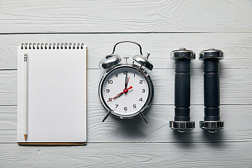 flat lay with silver alarm clock, empty notebook with pencil and dumbbells on wooden white background