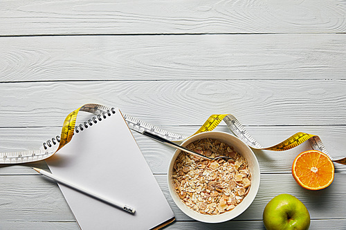 top view of notebook with pencil, breakfast cereal in bowl, apple and orange near measuring tape on wooden white background with copy space